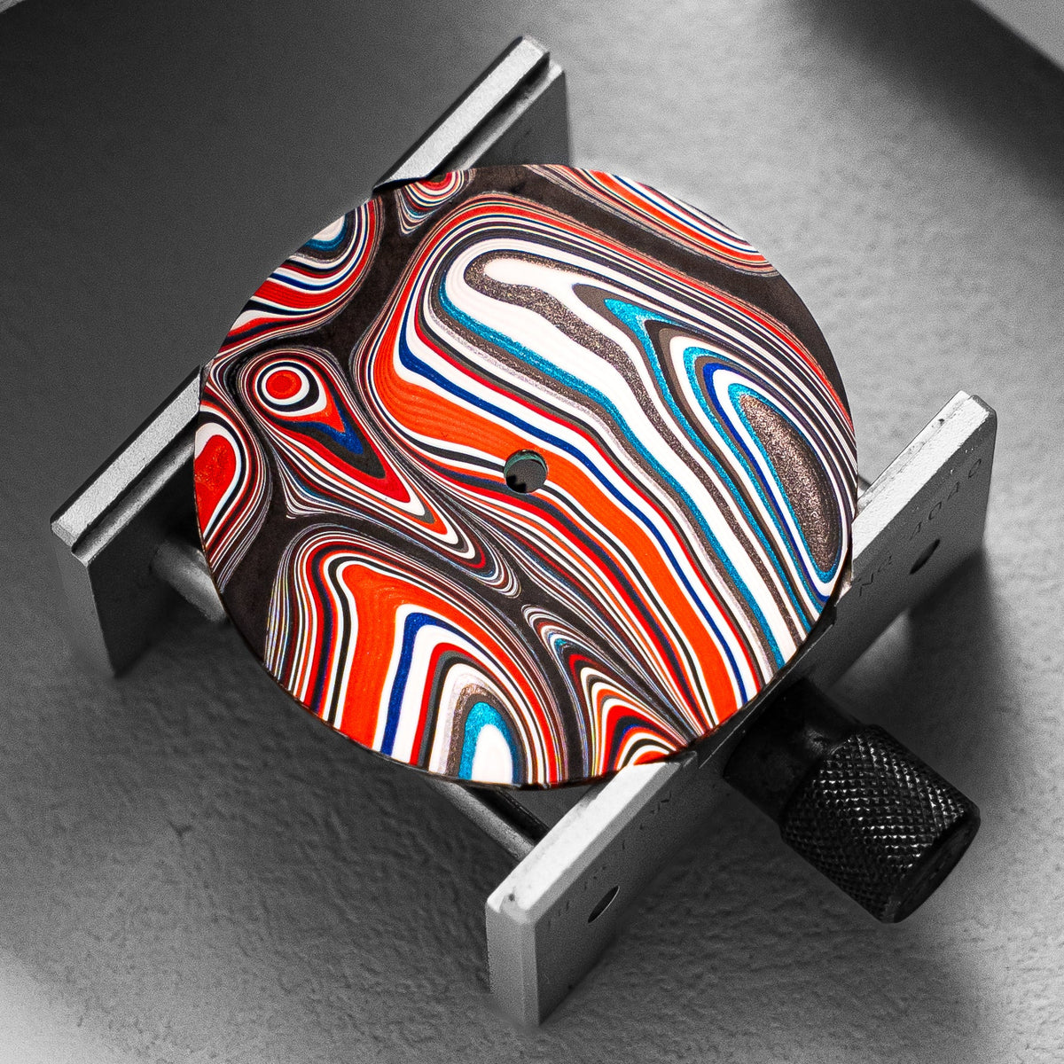 A close-up of a fordite watch dial with a swirling psychedelic pattern in red, orange, white, blue, and black, mounted on a silver metal bracket against a gray background.
Details